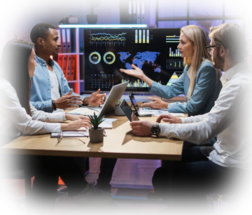 Four professionals sit around a conference table in a data analytics meeting, with a large screen showing a world map and charts behind them, and laptops on the table.